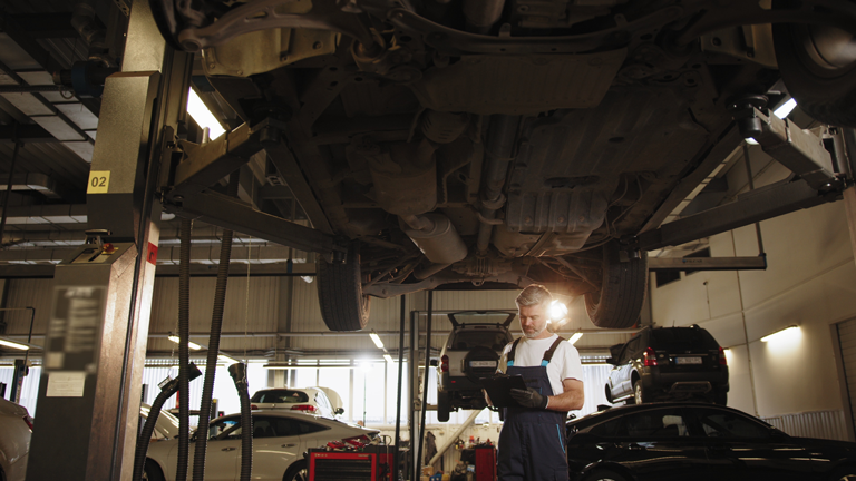 professional middle aged car mechanic taking notes under vehicle at auto repair shop